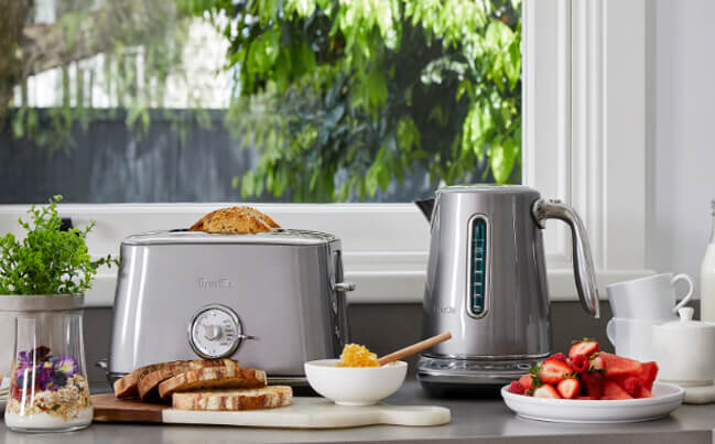 Silver toaster and kettle combo on a benchtop surrounded by bread and fruit