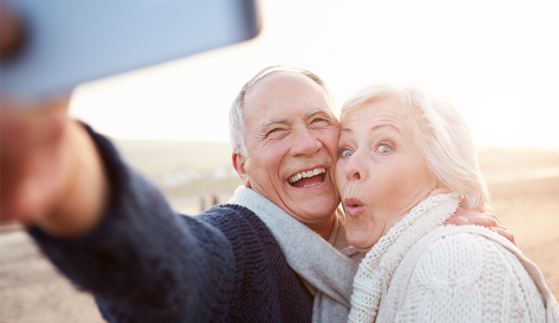 Lovely older couple are taking a selfie on the beach with their mobile phone. The husband is smiling and the wife is making a funny face.