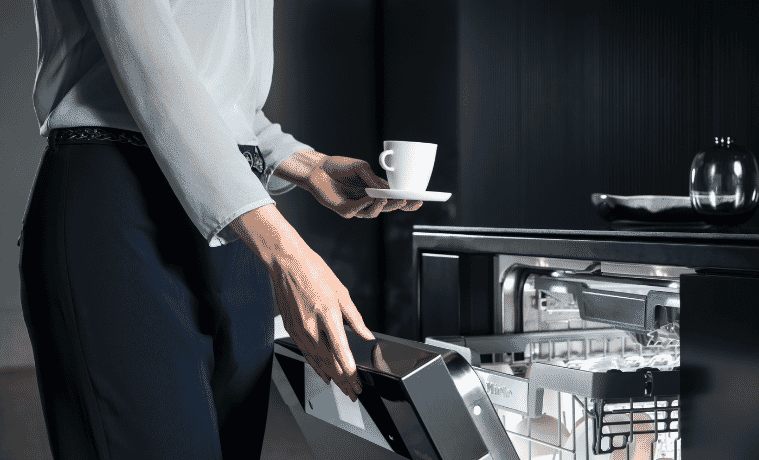 A woman opens her dishwasher to stack her empty cup and saucer.