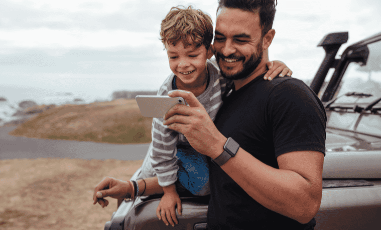 Dad and young son, standing next to their 4WD, near the coast, are both looking at a mobile phone and smiling.