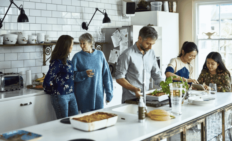 A man prepares home dinner for his extended family while they all gather around him in the kitchen to chat and socialise