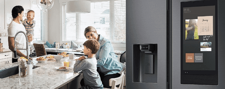 A Samsung Family Hub fridge sits off to the side of the kitchen of a young family having breakfast.