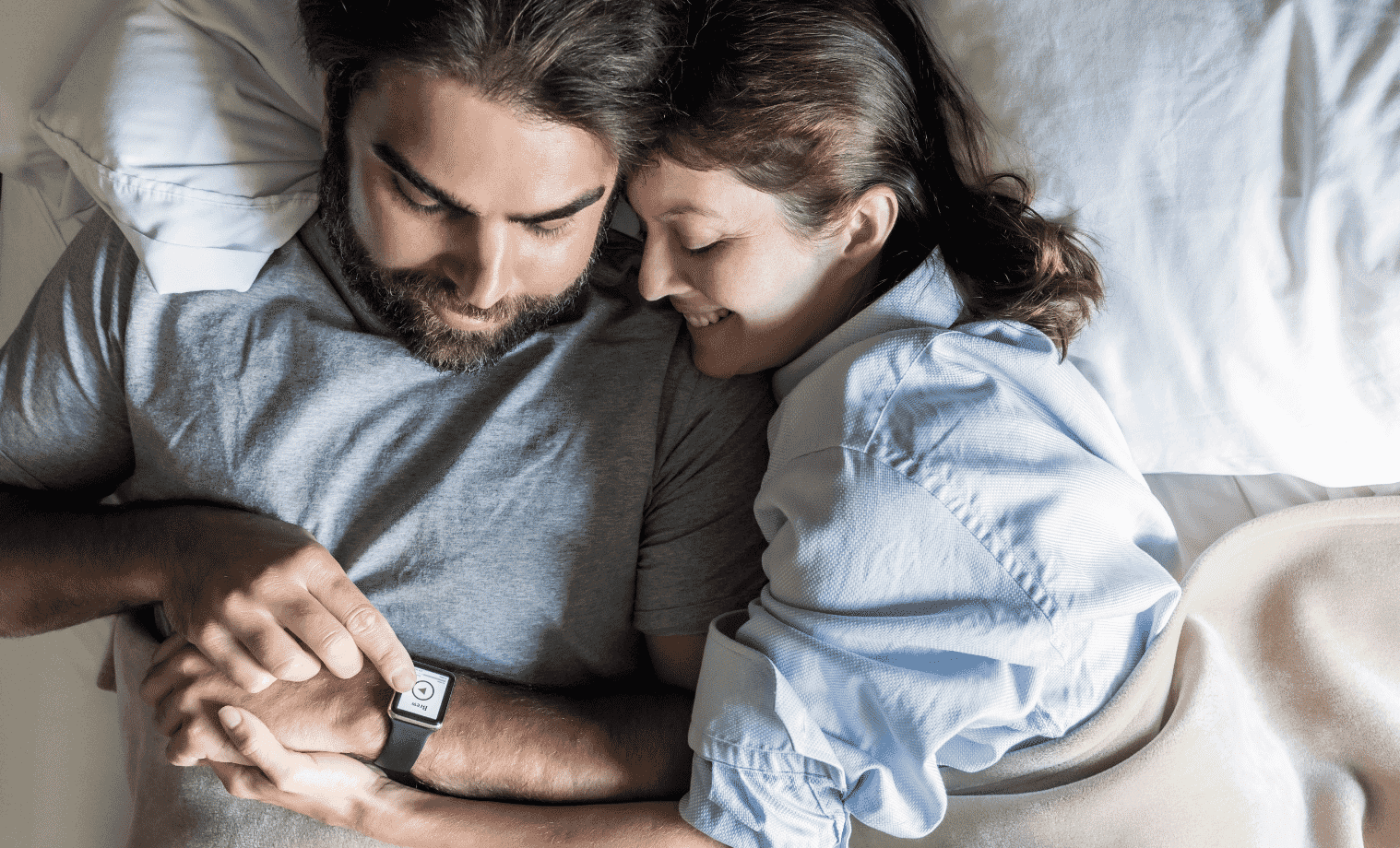 A man and woman lay in bed together and examine the man's smart watch.