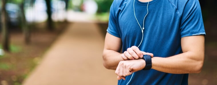 A man wears earphones and workout clothes in the park and pauses to check his fitness tracker.