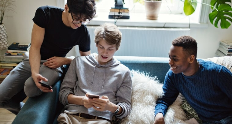 Three teenage sitting on a couch smiling whilst using mobile phones
