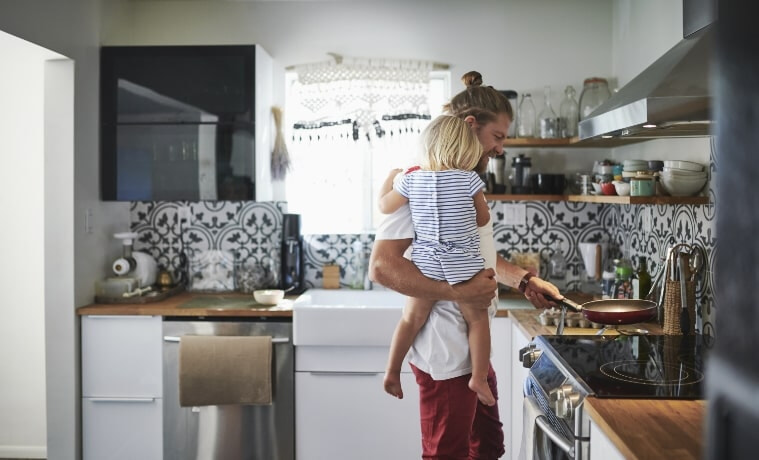 A father carrying his toddler daughter places an empty frying pan on an induction cooktop in his kitchen.