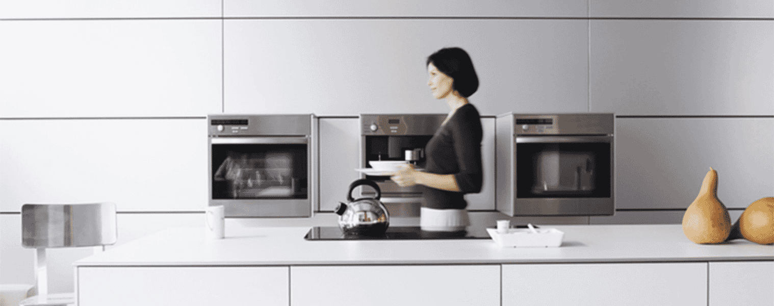 Woman walking through modern light coloured kitchen past a bank of ovens with pyrolytic cleaning.