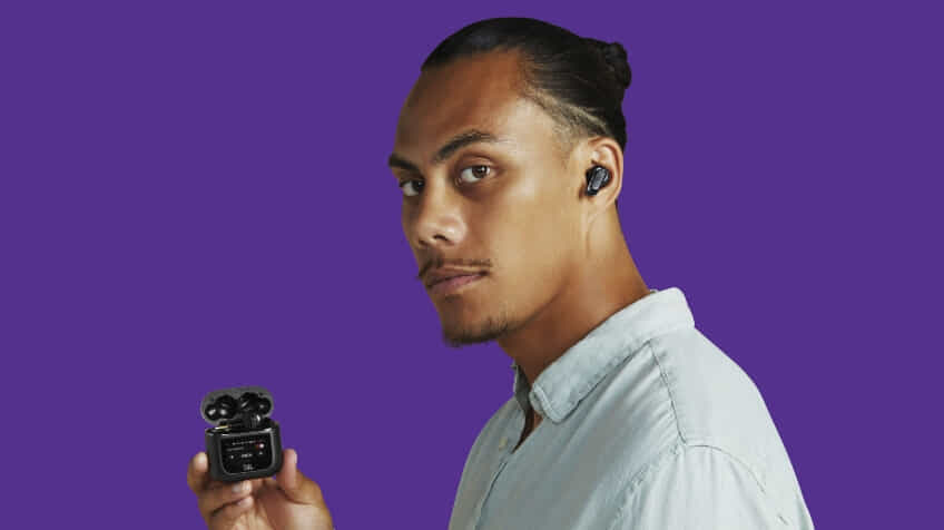 A man wears a pair of JBL True Wireless earbuds and holds the case on a purple background | The Good Guys