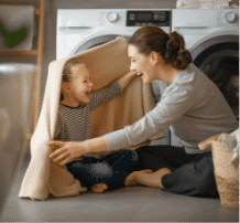 Mother and daughter playing under a blanket in the laundry in front of the washer and dryer.