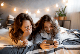 2 friends sitting on a bed laughing at something that appears on a phone.|The Good Guys 