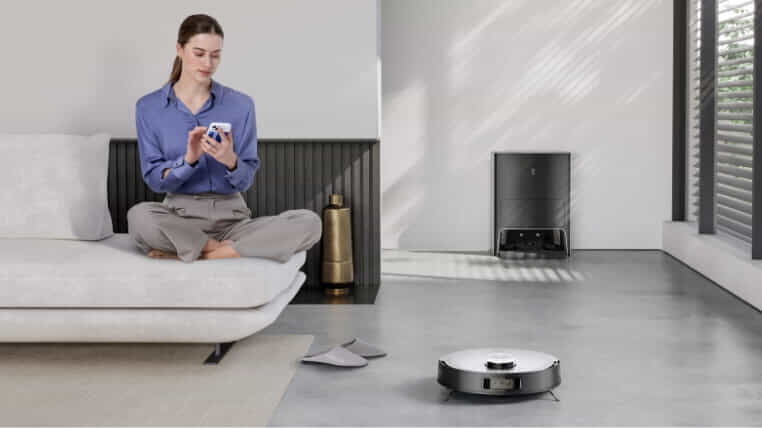 Woman sitting on a couch using her phone with her robot vacuum cleaning the floors in front of her.
