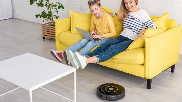 Mother and daughter sitting on a couch using a laptop with their feet up whilst the robot vacuum cleans underneath them.