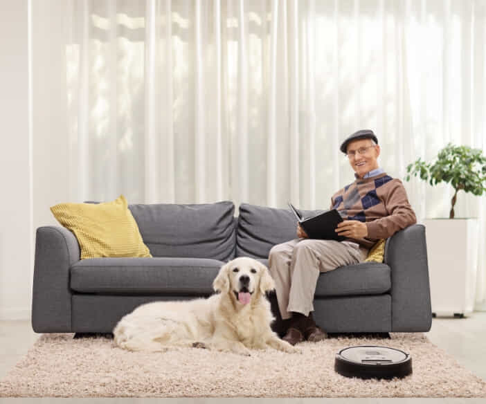 A man sitting on the couch with an iPad with his dog sitting on a rug on the floor in front of him whilst his robot vacuum cleans.