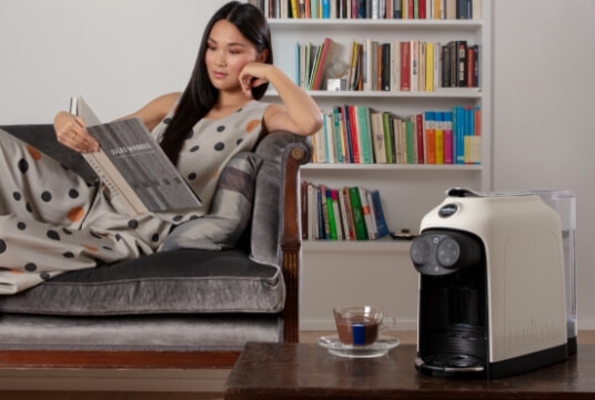 Person relaxing reading a book on the couch with their coffee machine to the right hand side of the image.