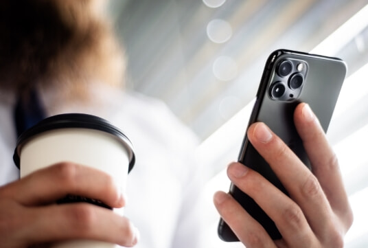 Close up of a person holding a mobile phone in one hand and a cup of coffee in the other.