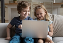 Two children smiling whilst holding a laptop on their laps.  