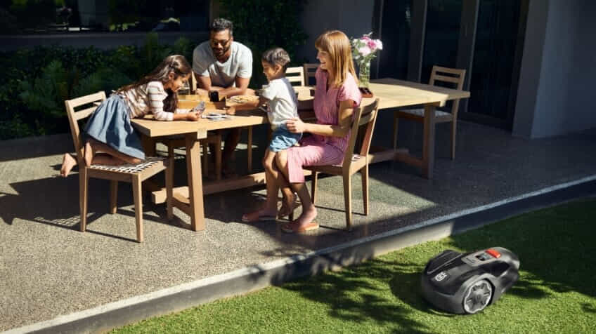 A young family have afternoon tea and play a card game while a Husqvarna robot lawn mower mows their lawn.