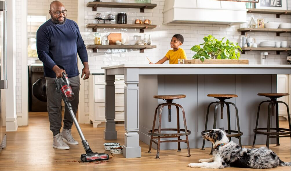 A father vacuums up his dog's spilled kibble while his young son watches.