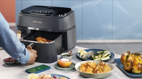 Image of someone using Philips 3000 Series Dual Basket Airfryer on benchtop next to various dishes of food.