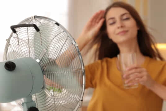 A woman drinks a glass of water while cooling herself in front of a fan.