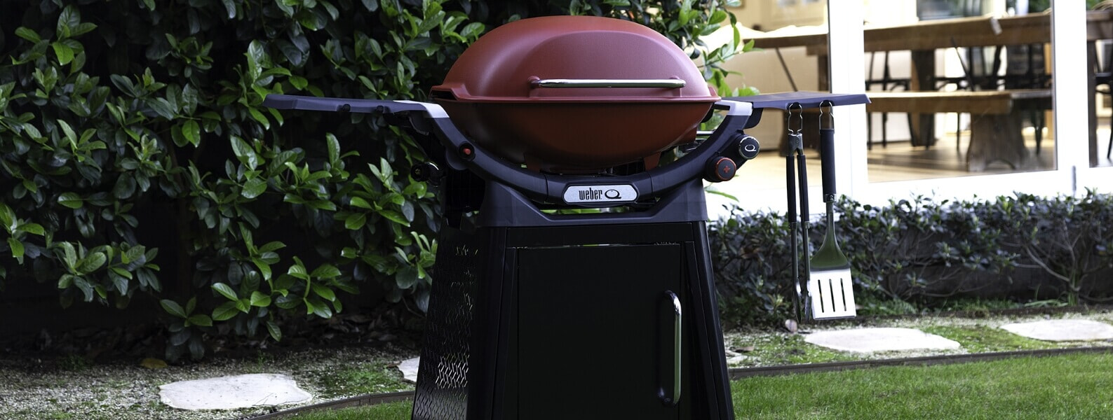 A black Weber Family Q BBQ stands next to an outdoor kitchen in a backyard | The Good Guys