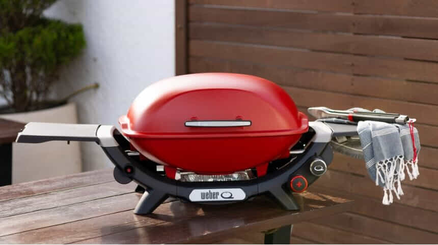 A red Weber Q+ BBQ sits on a table on a deck in a backyard.