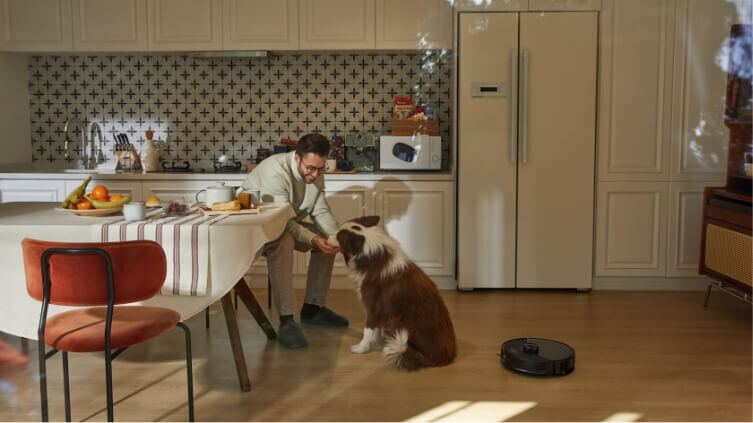 Man and dog in kitchen whilst robot vacuum cleans hard floors.