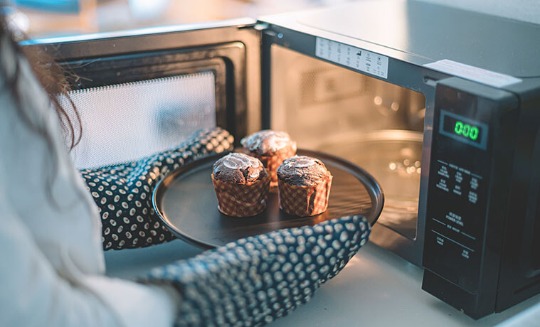 A woman with oven mitts pulling a tray of muffins out of a microwave.