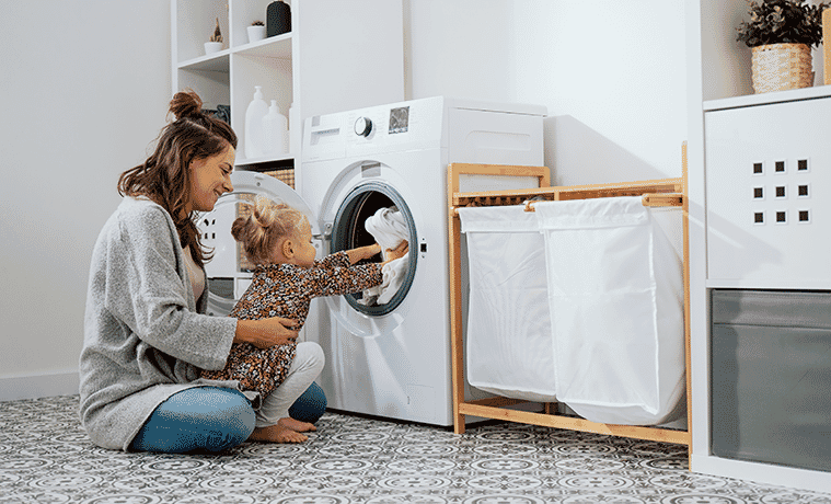 A mother and daughter sit on the laundry room floor to place clothes in the front loader washing machine.