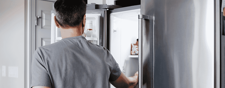 Man in a grey shirt opening the freezer door of a side-by-side fridge to look for ingredients.