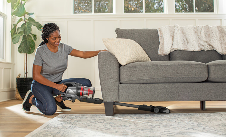 A woman vacuums under her couch with a Hoover stick vacuum.