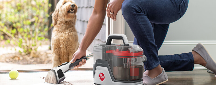 A woman uses her Hoover CleanSlate SpotWasher Carpet Cleaner to clean muddy pawprints off her entryway rug while her dog watches outside.