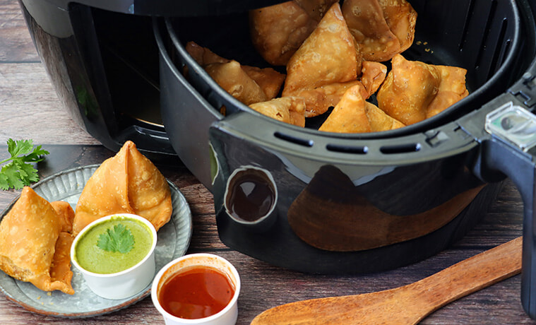 Close-up view of crunchy, golden samosas in an air fryer with a plate of samosas in the foreground.  