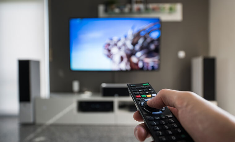 A man points his TV remote at a big screen TV mounted on the wall in his living room. 