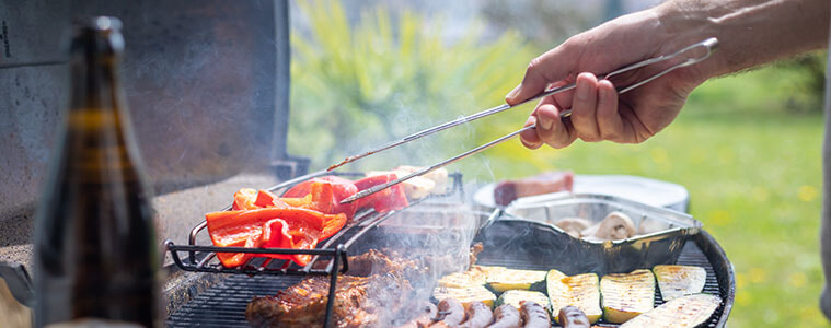 A man cooks sausages, steak and vegetables on an outdoor grill in his backyard.