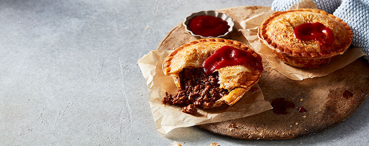 Crispy, golden homemade meat pies on a bench with a white cloth and a small dish of tomato sauce.