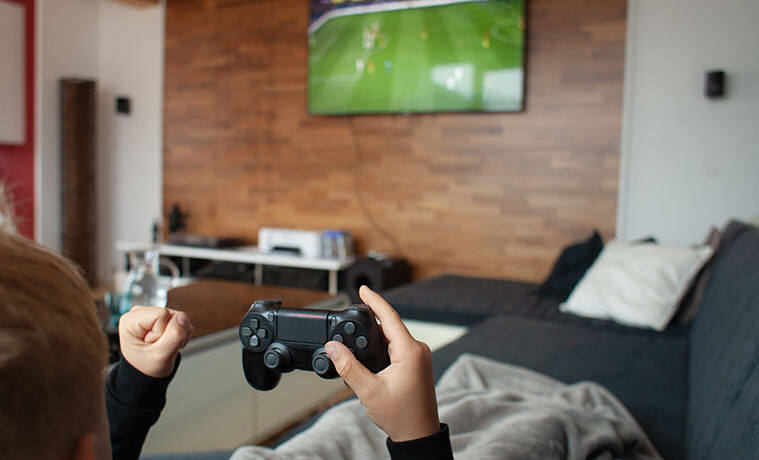 Over-the-shoulder view of a boy’s hands holding a gaming console in front of a TV screening a football game.