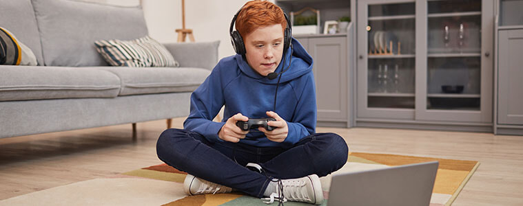Boy sitting on the floor of his living room wearing a gaming headset and holding his controller which is plugged into a laptop.