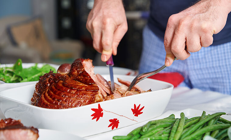 Closeup of a man using a knife and fork to carve a glazed ham at Christmas. 