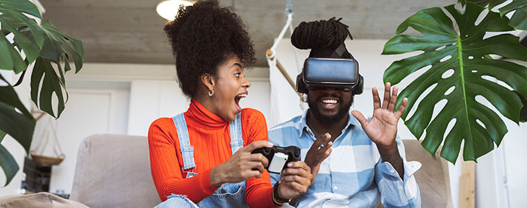 A young man and woman sit on the couch together, with the man wearing a VR headset to play a video game.