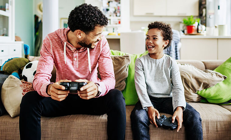A father and son hold gaming consoles and smile at each other while sitting on the couch during a video gaming session.