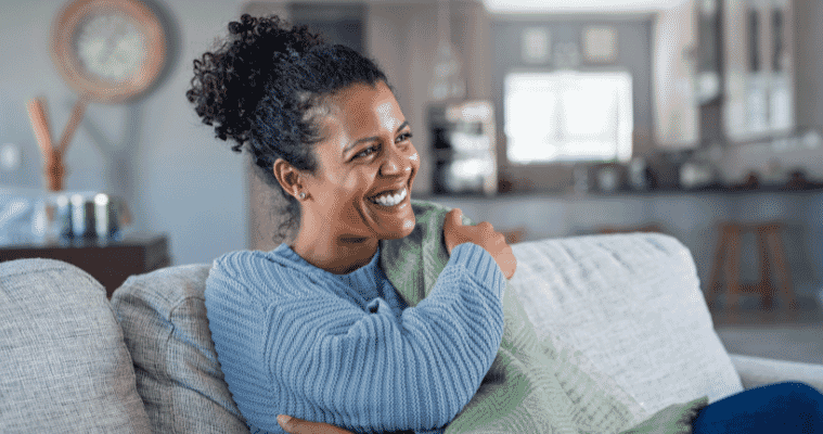 A woman on her couch underneath a blanker and a woman snuggling with a  dog. 