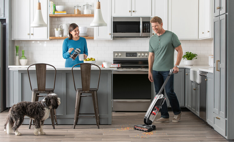A man vacuums his kitchen while his wife and dog watch.