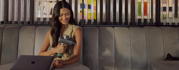 A woman sits at a table with her laptop and uses and Hyperice Hypervolt massage gun on her arm.