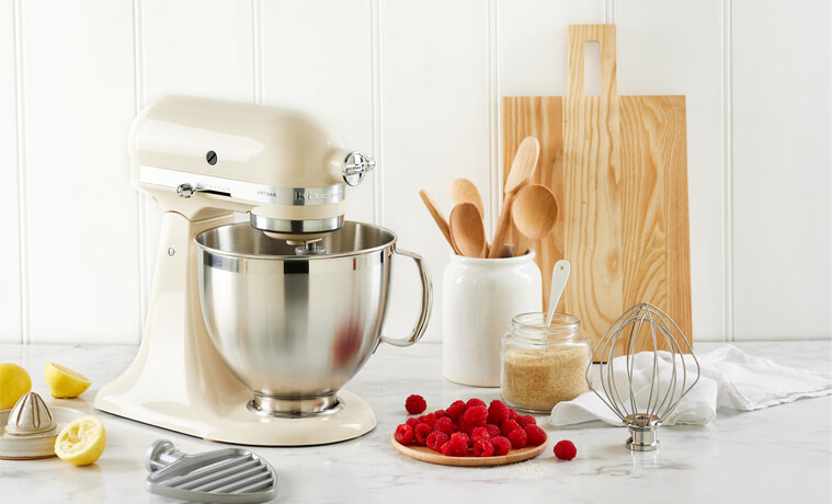 A cream coloured KitchenAid Artisan Stand Mixer sits in a white modern kitchen on a grey benchtop.