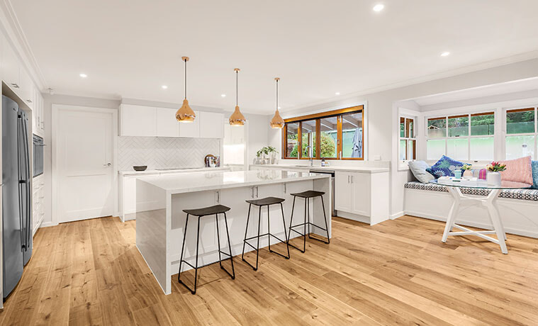 Bright open plan kitchen with black stools, timber floors and a trio of brass-look pendant lights above a white kitchen island.