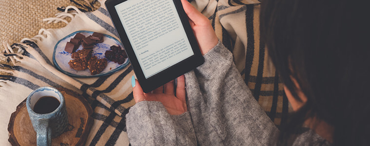 Woman reading a book on her ereader while wrapped in a warm blanket.  