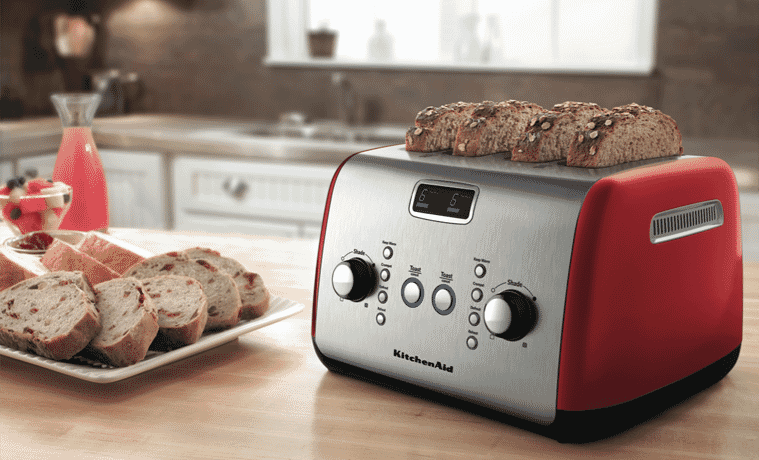 The KitchenAid Artisan 4 Slice Toaster in Empire Red sits on a butcher block kitchen benchtop with 4 slices of thick bread being toasted. A plate of toasted bread sits to the left of the toaster.