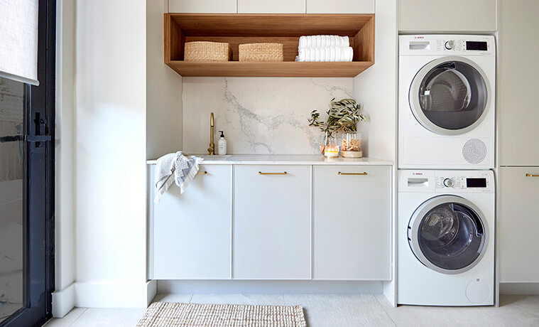 Modern laundry with white cabinetry, marble splashback, open timber shelf and stacked washing machine and dryer. 