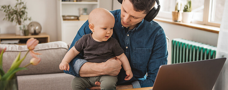 Young father wearing headphones works from home on his laptop, with his baby son on his lap.